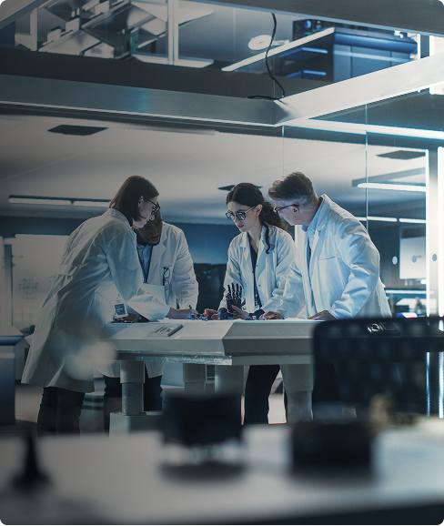 Quatro cientistas, vestidos com jalecos brancos, estão em um laboratório iluminado. Eles se curvam sobre uma bancada, analisando um projeto com equipamentos e objetos de pesquisa, em um ambiente de cocriação.